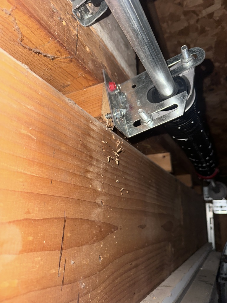 Torsion spring center bracket pulling away from a garage door header after its bottom lag screw stripped out, with wood chips below.