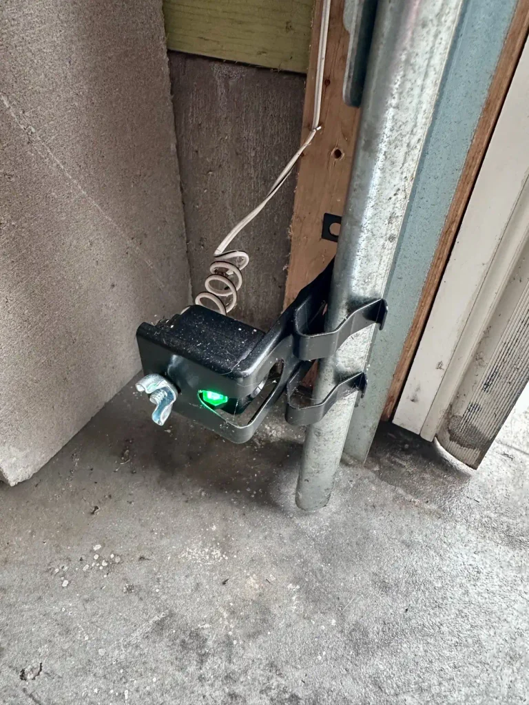 Garage door safety sensor with a green indicator light, mounted to the vertical track near the floor.